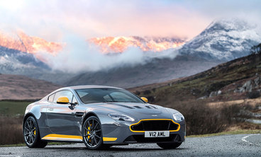Photo of an Aston Martin V12 Vantage S. It's gray with neon yellow details and sitting parked in front of a mountain with a sunset 